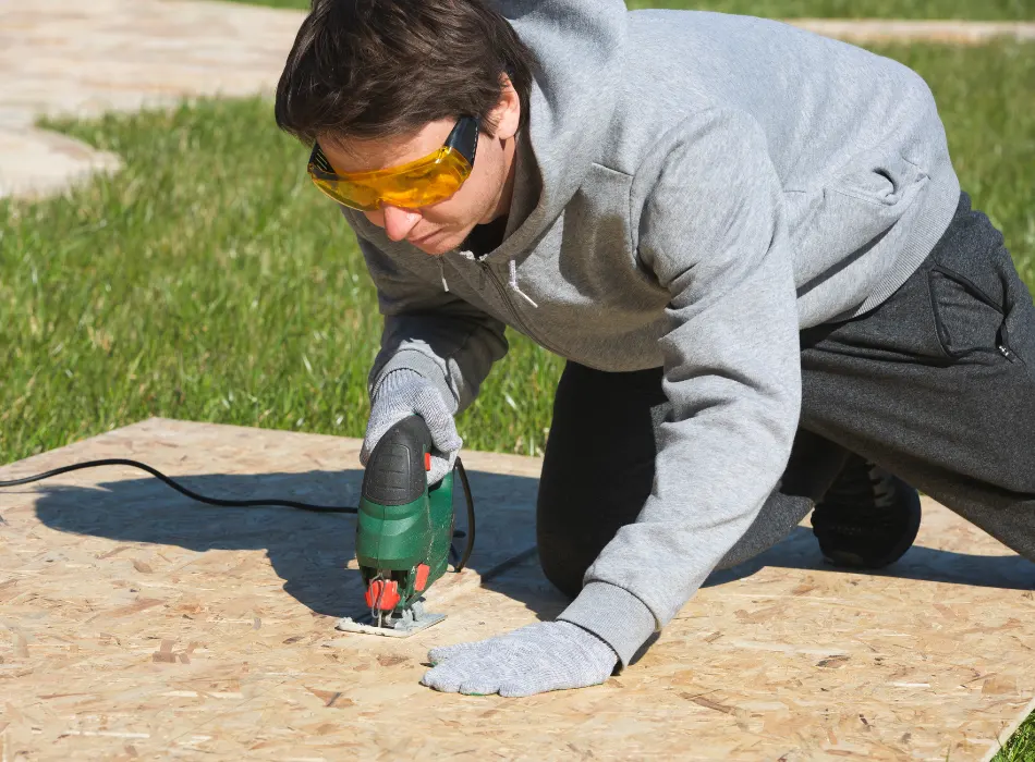 A man wearing goggles and using an electric saw to cut the OSB board